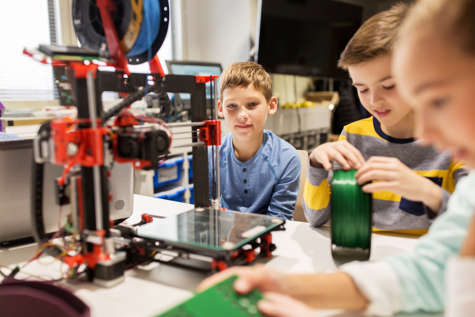Children gathered around a desktop 3D printer, handling filament and watching it print