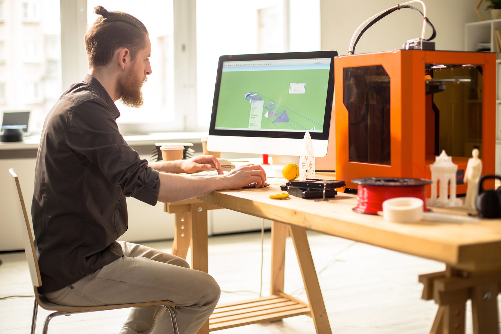 Person working at a desk with a computer and 3D printer in a home office setting.